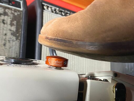 A shoe pressing a Barefoot Buttons V1 Standard Orange footswitch topper installed on a guitar pedal.