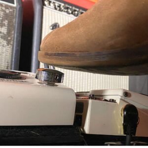 Barefoot Buttons V1 Standard Chrome footswitch topper installed on a white guitar pedal, pressed with a boot in front of guitar amps.